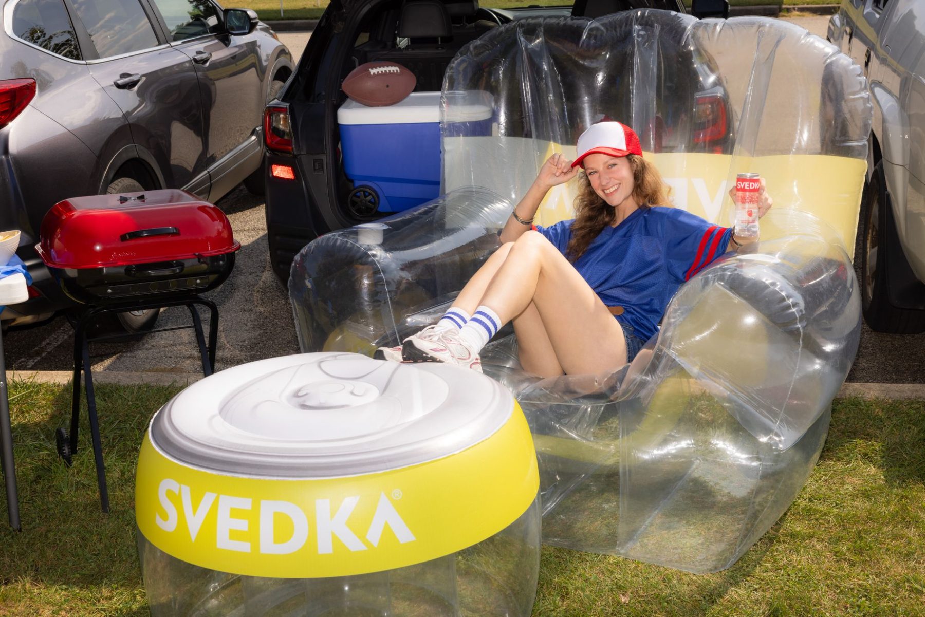 A tailgater lounges in the clear Pineapple Inflatable Tailgate Chair with her feet up on the yellow can-top footrest, holding a can of SVEDKA Vodka Water