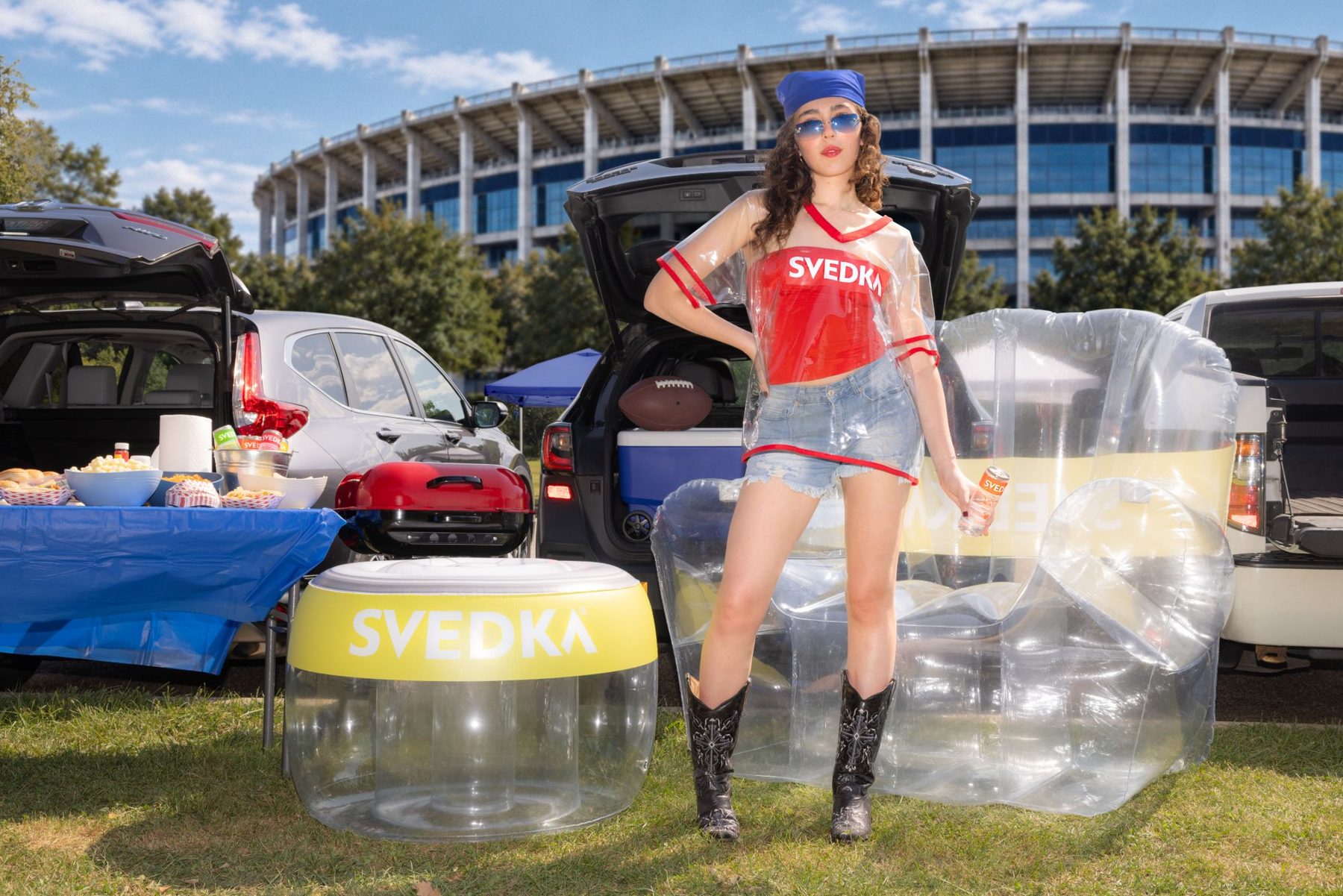 A tailgater in the Strawberry Oversized Jersey stands between the Pineapple Inflatable Tailgate Chair and its can-top footrest outside a stadium