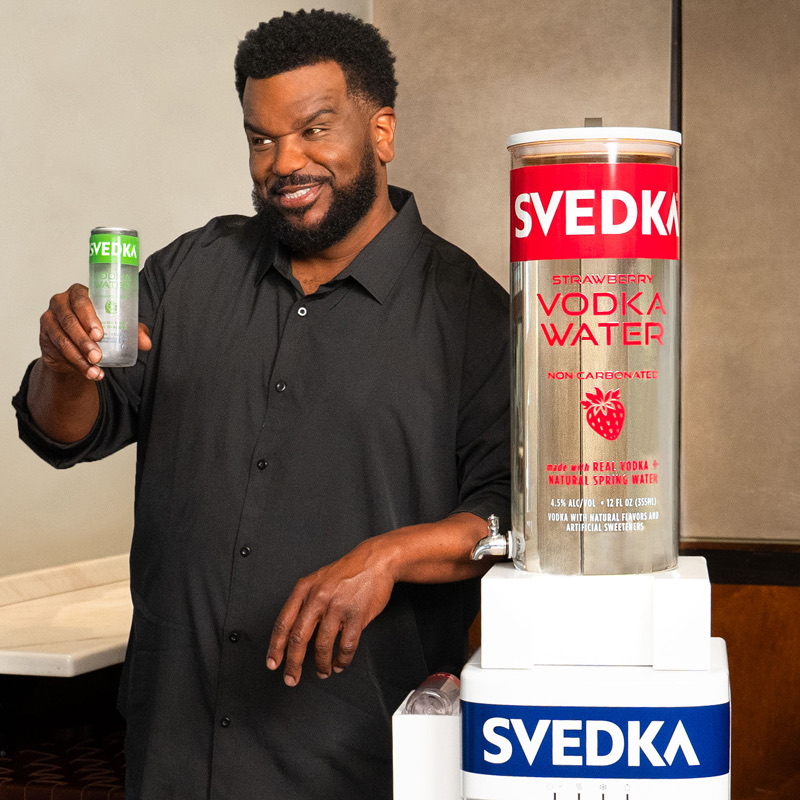 Craig Robinson smiling and holding a can of SVEDKA Lime Vodka Water next to the SVEDKA Water Cooler